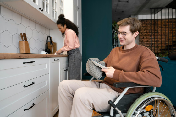 A young man in a wheelchair smiles while drying a dish with a towel in a modern kitchen. A woman in the background is washing dishes at the sink.