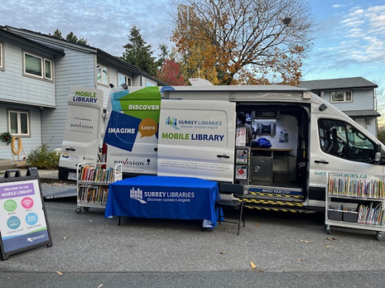 A Surrey Libraries Mobile Library van parked on a residential street. The side door is open, revealing the interior setup with books and equipment.