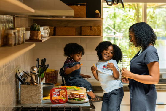 A smiling woman and two young children prepare food together in a bright kitchen. One child sits on the counter while the other stands beside the woman, all holding small vegetables.